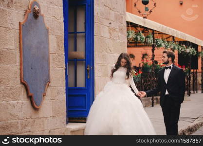 Wedding couple hugging in the old city. Blue vintage doors and cafe in ancient town on background. stylish bride in white long dress and groom in suit and bow tie. wedding day.. Wedding couple hugging in the old city. Blue vintage doors and cafe in ancient town on background. stylish bride in white long dress and groom in suit and bow tie. wedding day