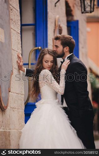 Wedding couple hugging in the old city. Blue vintage doors and cafe in ancient town on background. stylish bride in white long dress and groom in suit and bow tie. wedding day.. Wedding couple hugging in the old city. Blue vintage doors and cafe in ancient town on background. stylish bride in white long dress and groom in suit and bow tie. wedding day