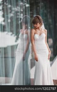Wedding day. Bride portrait in white dress by the window outside. Beauty portrait of bride wearing fashion wedding dress with luxury delight make-up and hairstyle.