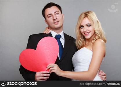 Wedding day. Happy blonde bride and groom holding red heart symbol gray background