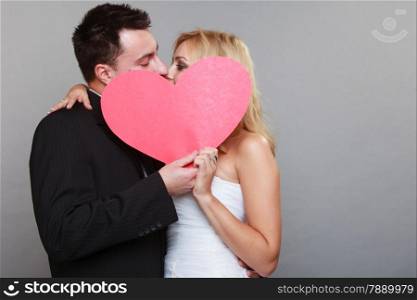 Wedding day. Happy blonde bride and groom kissing behind a red heart gray background