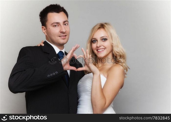 Wedding day. Happy blonde bride and groom showing making heart shape sign with hands gray background