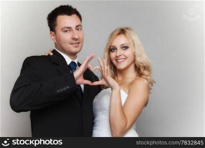 Wedding day. Happy blonde bride and groom showing making heart shape sign with hands gray background
