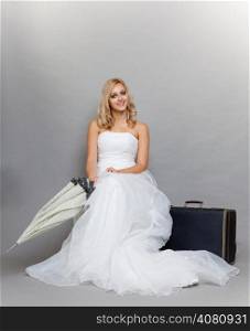 Wedding day. Portrait of beautiful young blonde bride wearing wedding dress in retro fashion style with umbrella and suitcase. Full length studio shot gray background