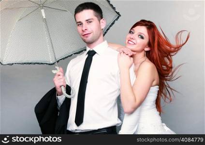 Wedding day. Portrait of happy married couple blue eyed bride with her red hair blowing in the wind and groom with umbrella studio shot on gray background