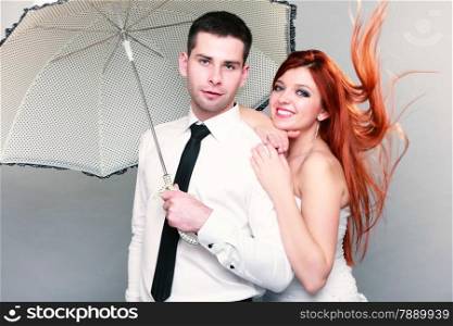 Wedding day. Portrait of happy married couple blue eyed bride with her red hair blowing in the wind and groom with umbrella studio shot on gray background