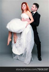 Wedding day. Portrait of happy married couple groom holding red haired bride up in his hands full length studio shot on gray background