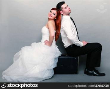 Wedding day. Portrait of happy married couple red haired bride and groom in full length sitting on old suitcase back on back studio shot on gray background. Honeymoon and travel