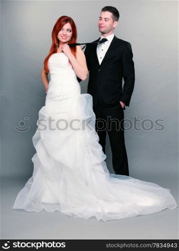 Wedding day. Portrait of happy married couple red haired bride and groom in full length. Woman pulling on mans tie, studio shot on gray background