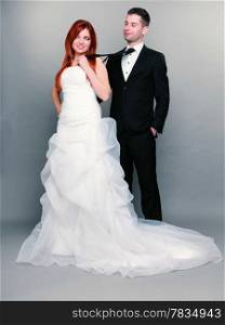 Wedding day. Portrait of happy married couple red haired bride and groom in full length. Woman pulling on mans tie, studio shot on gray background