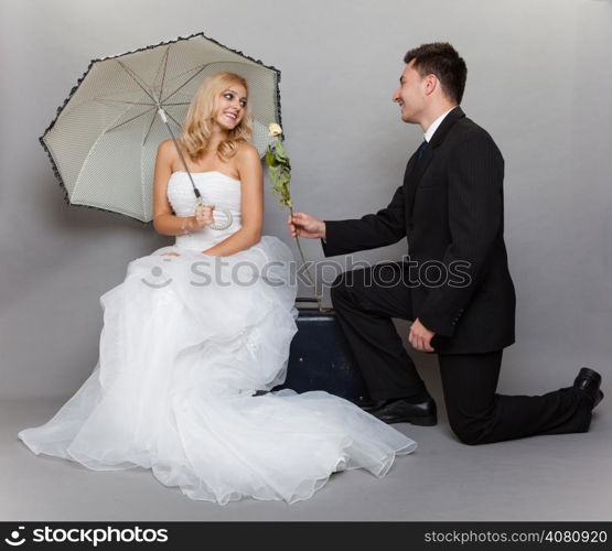 Wedding day. Portrait of romantic married couple blonde bride with umbrella and enamored groom giving a rose to girl. Full length studio shot gray background