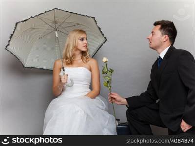 Wedding day. Portrait of romantic married couple blonde bride with umbrella and enamored groom giving a rose to girl. Studio shot gray background