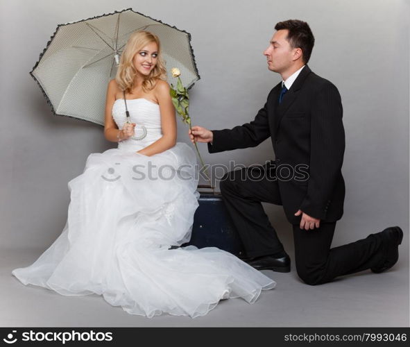 Wedding day. Portrait of romantic married couple blonde bride with umbrella and enamored groom giving a rose to girl. Full length studio shot gray background