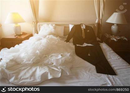 Wedding dress and suit lying on the white bed with beautiful sun light in hotel room. Valentines day and love for celebration concept.