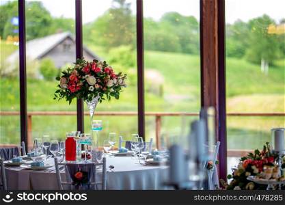 Wedding table decoration. Beautiful bouquet of flowers in vase on the table, next to plates, glasses and gift boxes. Bouquet of flowers, glasses, gift boxes and jugs setting on the festive table in restaurant.