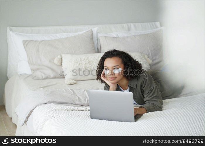 Weekend in bed. Lovely afro girl applies eye patches and relaxing at home. Girl is watching movie on laptop. Daily beauty routine of teenage girl. Cosmetic applying, hygiene and dermatology.. Weekend in bed. Lovely afro girl applies eye patches and relaxing at home watching movie.