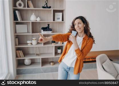 Weekend morning at home. Emotional caucasian woman is singing with phone as with microphone. Carefree girl having fun in her living room in modern apartment. Electronic gadgets using.. Weekend morning at home. Emotional caucasian woman is singing with phone as with microphone.