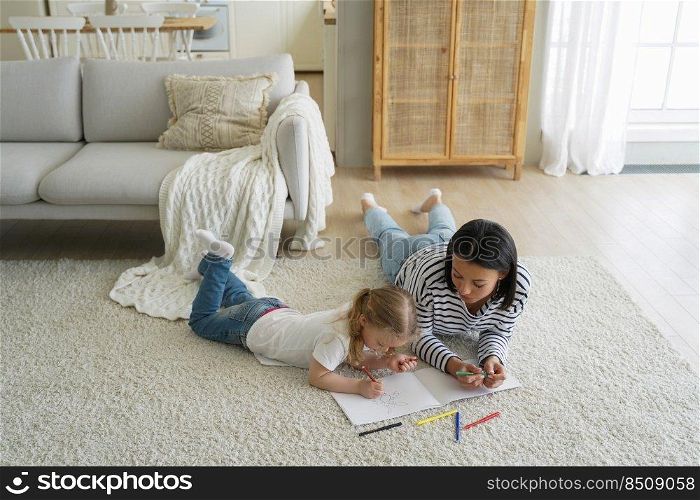 Weekend morning of the family, mum and toddler. Woman and little girl relaxing, lying on floor together and painting with colorful markers. European mum teaching her kid how to draw in coloring book.. Weekend morning of family. Mum and kid lying on floor together and painting with colorful markers.