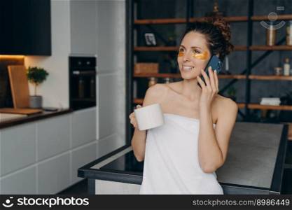Weekend morning phone talk. Young woman applies anti wrinkle eye patches and relaxing at home talking on smartphone. Happy hispanic girl wrapped in towel after skincare procedures. Modern interior.. Weekend morning phone talk. Young woman applies anti wrinkle eye patches and talking on phone.