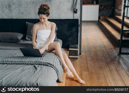 Weekend of a freelancer. Attractive european woman wrapped in towel after bathing is sitting at laptop on her bed. Young woman takes shower and relaxing at home. Remote worker lifestyle.. Weekend of a freelancer. Young woman wrapped in towel after bathing is sitting at laptop on her bed.
