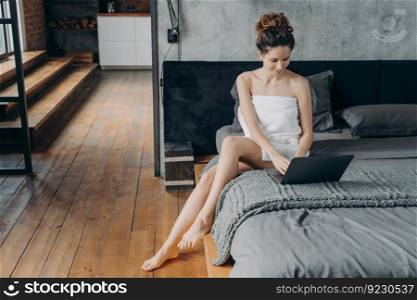 Weekend of a freelancer. Attractive european woman wrapped in towel after bathing is sitting at laptop on her bed. Young woman takes shower and relaxing at home. Remote worker lifestyle.. Weekend of a freelancer. Young woman wrapped in towel after bathing is sitting at laptop on her bed.