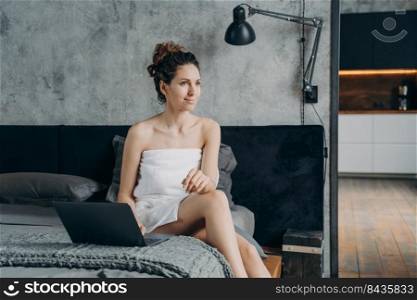 Weekend of a freelancer. Happy girl works through internet. Young attractive european woman wrapped in towel after bathing is sitting at laptop on her bed. Worker spends vacation in hotel.. Weekend of a freelancer. Young attractive european woman works through internet in hotel.