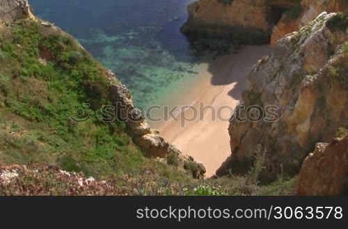 Wei?er verlassener Natursandstrand vor turkisblauem Wasser zwischen hohen, steilen, teilweise grun bewachsenen sandsteinfarbenen Felsen / Klippen - Kuste der Algarve, Portugal.