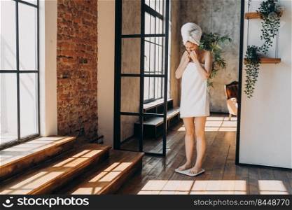Weight gaining concept. Frustrated slim european woman taking step onto scale to measure her weight. Girl wrapped in towel after bathing has time at spa. Stressed woman checking weight after shower.. Weight gaining concept. Frustrated slim european woman taking step onto scale to measure her weight.