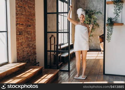 Weight loss concept. Excited European woman weighs herself on sca≤. Towel-wrapped girl enjoys spa time post-bath. Overjoyed woman checks weight after shower.