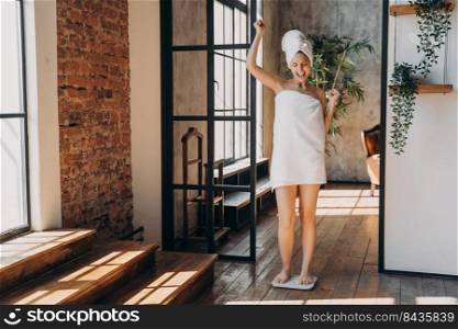 Weight loss concept. Excited slim european woman taking step onto scale to measure her weight. Girl wrapped in towel after bathing has time at spa. Overjoyed woman checking weight after shower.. Weight loss concept. Excited slim european woman taking step onto scale to measure her weight.