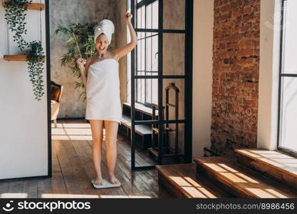 Weight loss concept. Excited slim european woman taking step onto scale to measure her weight. Girl wrapped in towel after bathing has time at spa. Overjoyed woman checking weight after shower.. Weight loss concept. Excited slim european woman taking step onto scale to measure her weight.