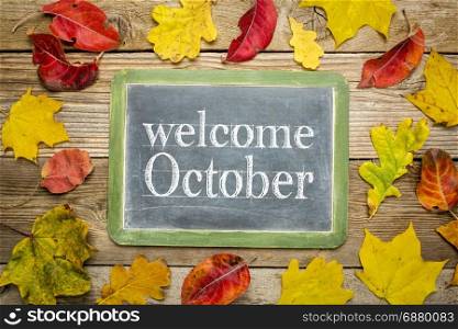 Welcome October on a slate blackboard against rustic weathered wood planks with colorful dried leaves