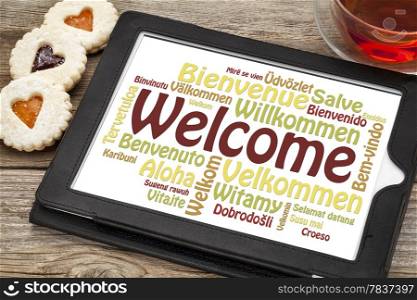 welcome word cloud in different languages on a digital tablet with a cup of tea and heart cookies