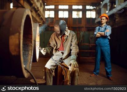 Welder in mask works with big metal pipe on factory, welding skill, worker in helmet on background. Metalworking industry, industrial manufacturing of steel products. Welder works on factory, worker on background
