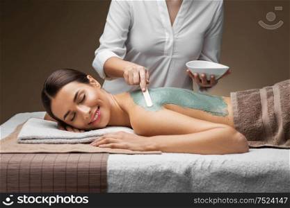 wellness, beauty and cosmetology concept - beautiful young woman lying with closed eyes and cosmetologist applying clay mask by spatula at spa. woman and cosmetologist applying clay mask in spa