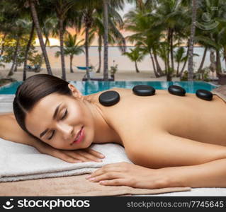 wellness, beauty and relaxation concept - beautiful young woman having hot stone massage at spa over tropical beach background in french polynesia. woman having hot stone massage at spa