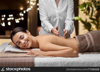 wellness, beauty and relaxation concept - beautiful young woman lying and having back massage at spa over christmas lights on window background. woman lying and having back massage at spa