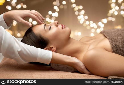 wellness, beauty and relaxation concept - beautiful young woman lying with closed eyes and having face and head massage at spa over festive lights on background. woman having face and head massage at spa