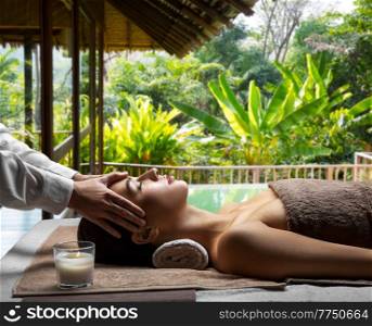wellness, beauty and relaxation concept - beautiful young woman lying with closed eyes and having face and head massage at spa over bungalow at exotic resort in thailand on background. woman having face and head massage at spa