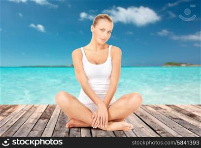 wellness, health and people concept - beautiful young woman in cotton underwear sitting on wooden floor over sea and blue sky background