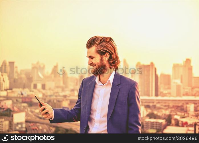 Western businessman using a phone on a rooftop with blurry city . Western businessman using a phone on a rooftop with blurry city background. Western businessman using a phone on a rooftop with blurry city background