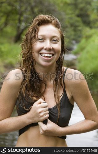Wet Caucasian young adult woman standing in stream wringing water out of her shirt smiling and looking at viewer.