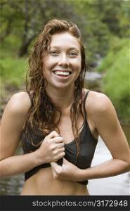Wet Caucasian young adult woman standing in stream wringing water out of her shirt smiling and looking at viewer.