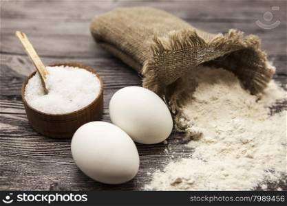 Wheat flour in a canvas bag, a large salt shaker wood, raw eggs: set for making homemade bread dough on a beautiful dark wooden background.