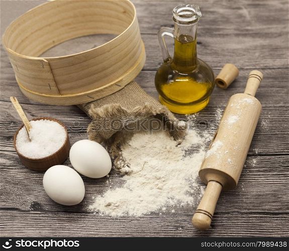 Wheat flour in a canvas bag,sieve, the olive oil in a glass carafe, a large salt shaker wood, raw eggs, a wooden rolling pin: set for making homemade bread dough on a beautiful dark wooden background.