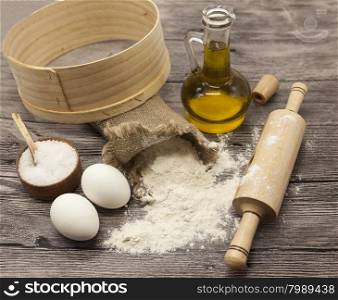 Wheat flour in a canvas bag,sieve, the olive oil in a glass carafe, a large salt shaker wood, raw eggs, a wooden rolling pin: set for making homemade bread dough on a beautiful dark wooden background.