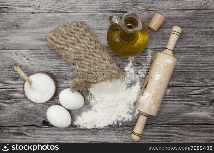 Wheat flour in a canvas bag, the olive oil in a glass carafe, a large salt shaker wood, raw eggs, a wooden rolling pin: set for making homemade bread dough on a beautiful dark wooden background.