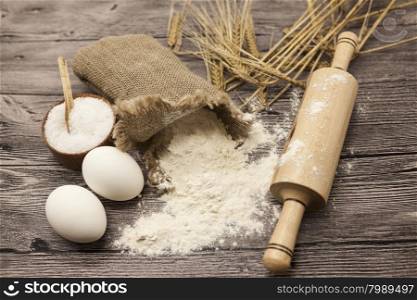 Wheat flour in a canvas bag, with spikelets of rye, a large salt shaker wood, raw eggs, a wooden rolling pin: set for making homemade bread dough on a beautiful dark wooden background.
