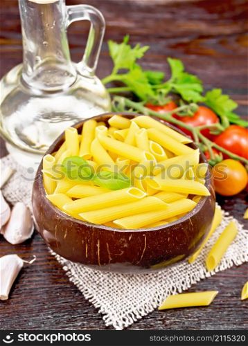 Wheat flour penne pasta in a bowl of coconut shells on sacking, tomatoes, garlic, vegetable oil in a glass decanter and parsley on dark wooden board background