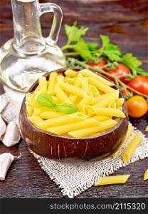 Wheat flour penne pasta in a bowl of coconut shells on sacking, tomatoes, garlic, vegetable oil in a glass decanter and parsley on dark wooden board background